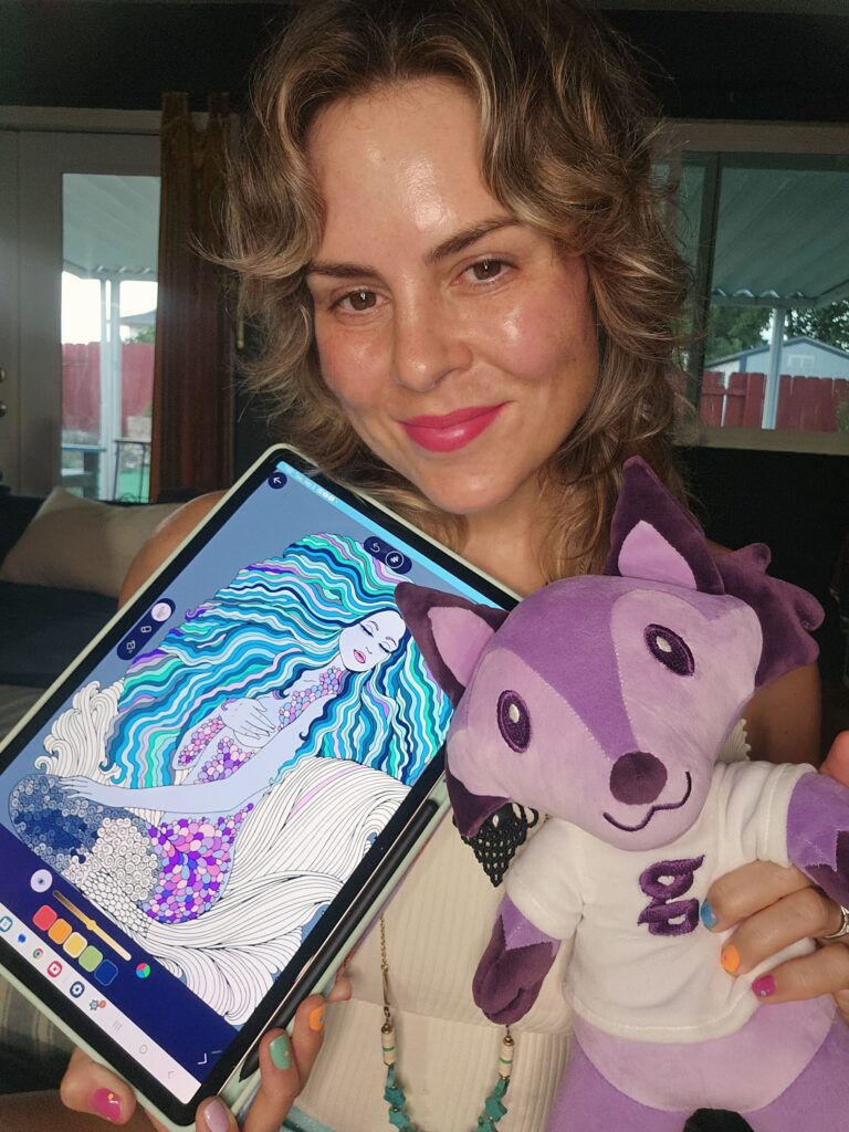 An image of Thera Barnett, the creator of the Groovy Coloring, an adult coloring book app. She is holding her tablet with the app on it and a plushie.